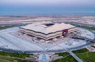 Estádio em formato de tenda em área árida, com canteiro de obras e estradas. Vista aérea.