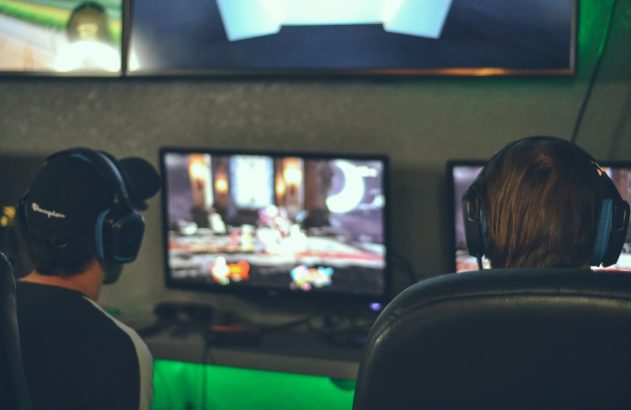 Na imagem é possível ver, de costas, duas pessoas jogando criptogames em monitores. As duas pessoas estão sentadas em cadeiras e usando headsets. Uma das pessoas usa boné.