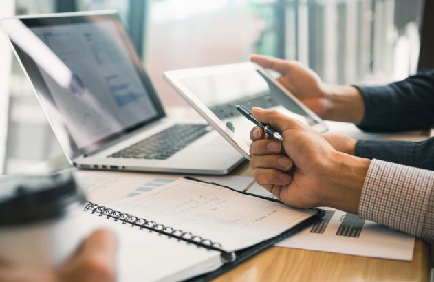 A imagem contém as mãos de duas pessoas brancas. A pessoa mais ao fundo segura um tablet, enquanto a mais próxima da câmera aponta para o mesmo com uma caneta na mão, enquanto segura um copo na outra. Na mesa, há um notebook prateado, uma agenda aberta, alguns papéis com gráficos impressos.