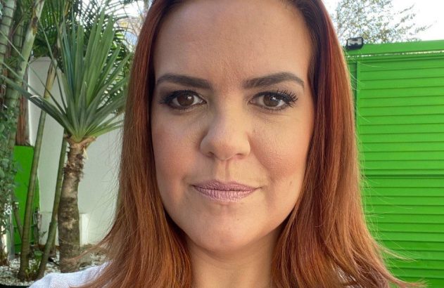 Mulher ruiva olhando para câmera, com colar de coração. Fundo com plantas e parede verde.