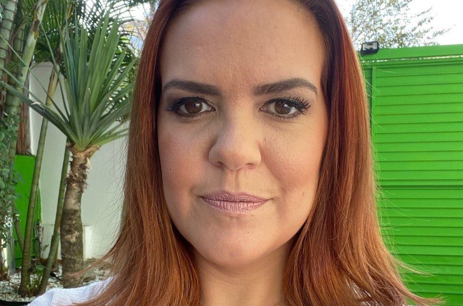 Mulher ruiva olhando para câmera, com colar de coração. Fundo com plantas e parede verde.