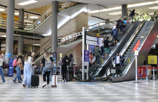 Interior de aeroporto com pessoas em escadas rolantes e caminhando com bagagens.
