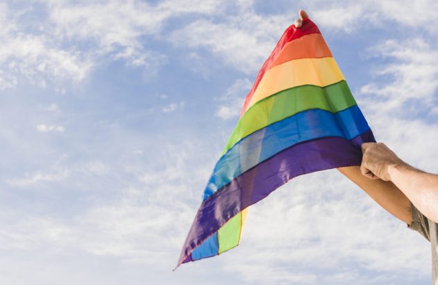 Pessoa segura bandeira arco-íris com cores vibrantes contra céu azul e nuvens.