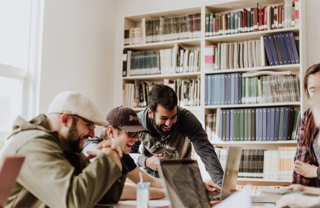 Grupo de amigos rindo e trabalhando juntos em laptops em uma biblioteca. Prateleiras cheias de livros ao fundo.