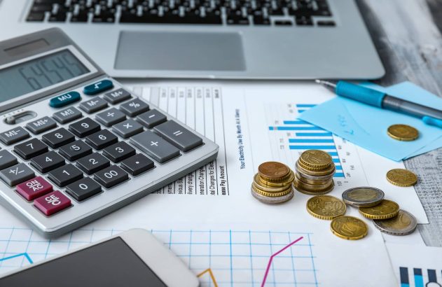 A imagem mostra uma notebook prateado com teclas pretas, uma calculadora, uma caneta azul, algumas moedas e um celular branco. Todos esses objetos estão dispostos em cima de papéis com gráficos e outras informações.