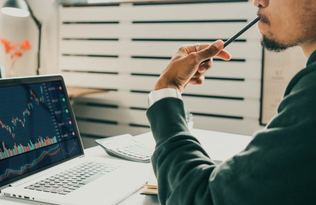 A imagem mostra um homem negro no que parece ser um escritório coletivo. Ele segura um lápis preto próximo a sua boca e sua mesa contém uma calculadora e um notebook, ambos na cor branca. Na tela do notebook, há gráficos de linha e de barra nas cores verde e vermelho, exemplificando o risco de liquidez.