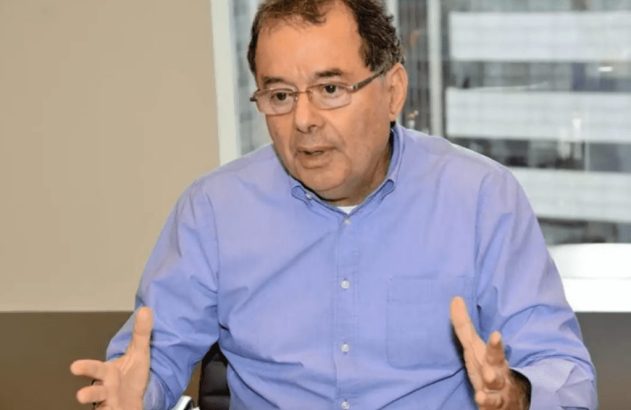 Homem de óculos e camisa azul, sentado, gesticula com as mãos abertas.