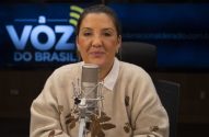 Mulher sentada à frente de microfone em estúdio de rádio com "A Voz do Brasil" na tela.