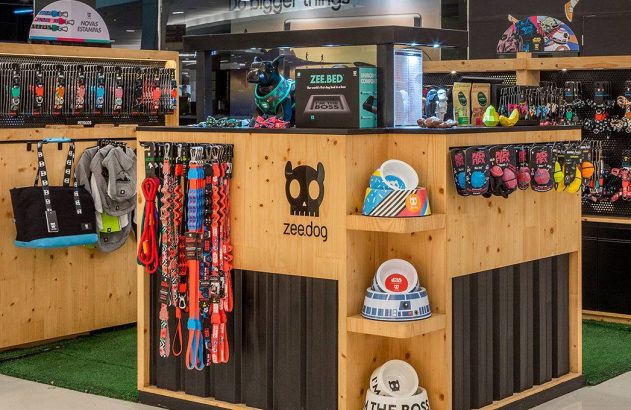 Expositor da Zee.Dog em madeira e preto, exibindo coleiras, guias, potes, camas e brinquedos para pets.