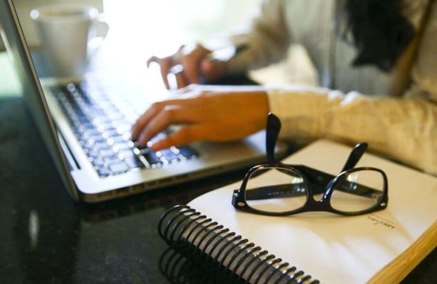 Mãos digitando em laptop. Óculos e caderno em espiral na mesa.