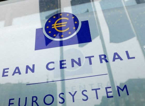 Sinal do Eurosystem com logo do euro (€) em vidro. Reflexos de bandeiras e edifícios.