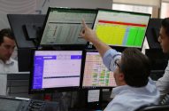 Dois homens em um ambiente de trabalho digital com múltiplos monitores, um deles aponta para a tela com dados financeiros.