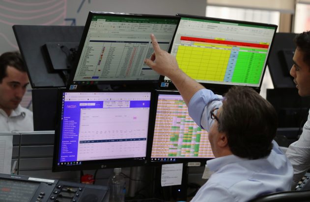 Dois homens em um ambiente de trabalho digital com múltiplos monitores, um deles aponta para a tela com dados financeiros.