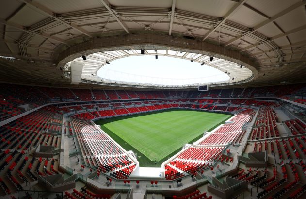 Estádio de futebol vazio, com campo verde e assentos vermelhos e pretos. Teto com estrutura aparente e abertura circular.