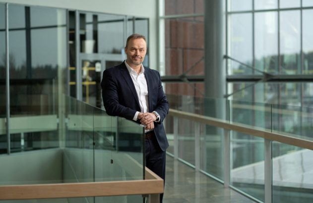 Homem de terno escuro, apoiado em corrimão de vidro em hall moderno, olha para a câmera.