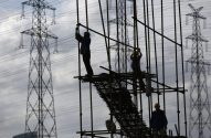 Trabalhadores da construção civil em andaimes, silhuetados contra torres de transmissão elétrica e céu nublado.