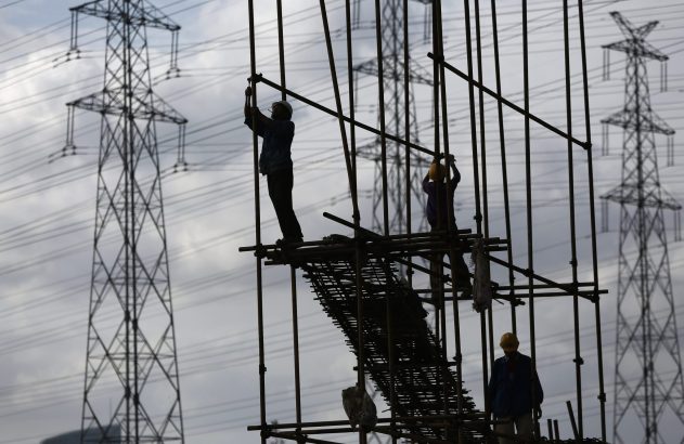 Trabalhadores da construção civil em andaimes, silhuetados contra torres de transmissão elétrica e céu nublado.