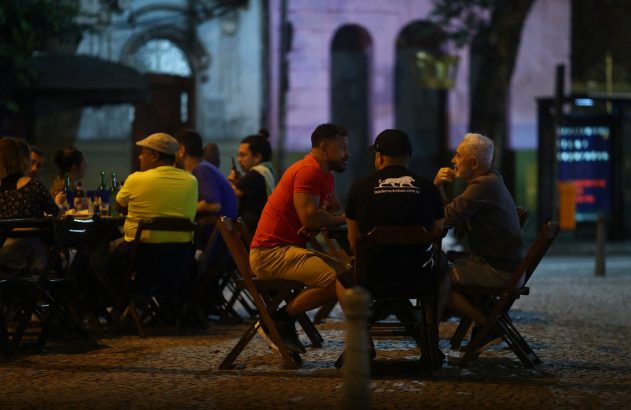 Pessoas sentadas em mesas ao ar livre à noite, conversando e bebendo em um ambiente de bar.