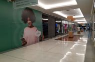 Corredor iluminado de shopping com painel verde de mulher sorrindo e placa de direções para lojas e lazer.