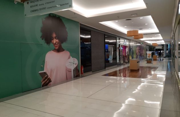 Corredor iluminado de shopping com painel verde de mulher sorrindo e placa de direções para lojas e lazer.