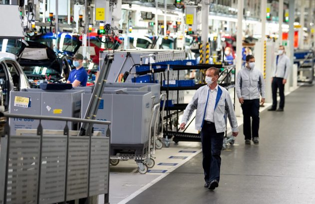 Homem de máscara caminha em linha de montagem de carros com outros trabalhadores e veículos em produção.