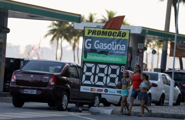 Mulher e duas crianças caminham em posto de gasolina. Letreiro de preços de gasolina à vista, com carros estacionados.