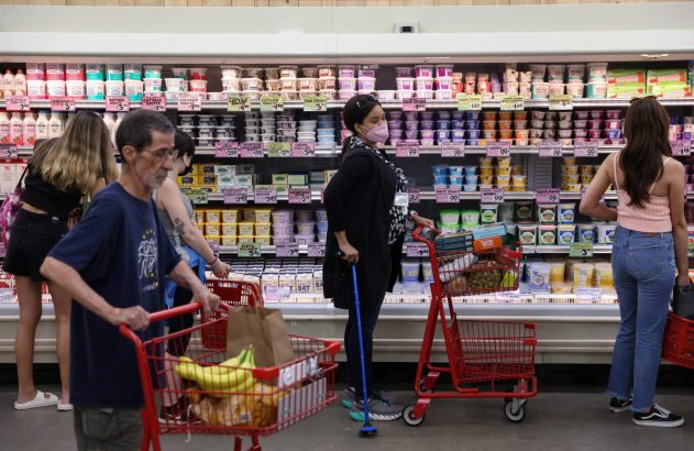 Pessoas fazendo compras em um supermercado, olhando produtos na seção de laticínios refrigerados. Há carrinhos vermelhos.
