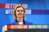 Liz Truss, séria, em blazer azul, em frente a um painel azul com o texto 'GETTING ON WITH THE JOB'.
