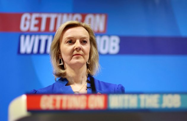 Liz Truss, séria, em blazer azul, em frente a um painel azul com o texto 'GETTING ON WITH THE JOB'.