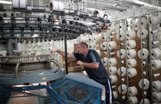 Homem em fábrica têxtil ajusta máquina de tricô circular. Centenas de cones de fio branco ao redor.