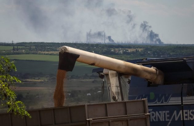Colheitadeira descarrega grãos em caminhão. Fumaça escura sobe de complexo industrial ao longe sobre paisagem rural.