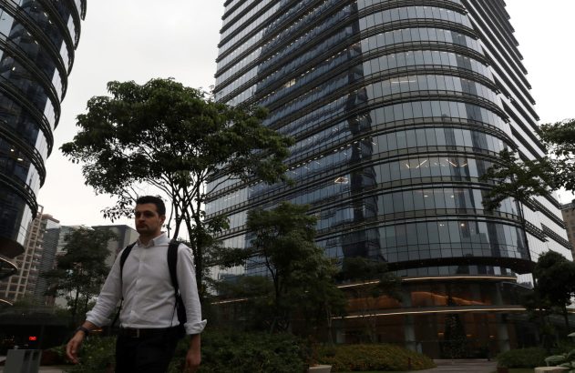 Homem de camisa branca e mochila caminhando em paisagem urbana com modernos edifícios de vidro e árvores.