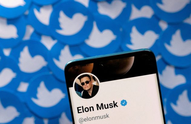 Um smartphone exibe o perfil verificado de Elon Musk no Twitter, com vários logos azuis da plataforma no fundo desfocado.