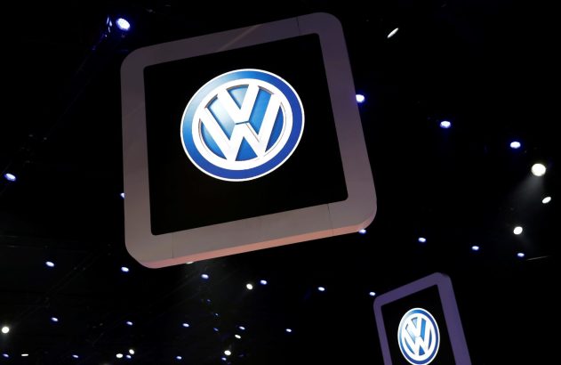 Logotipo da Volkswagen iluminado em display suspenso contra fundo escuro com pontos de luz; outro logo parcialmente visível.