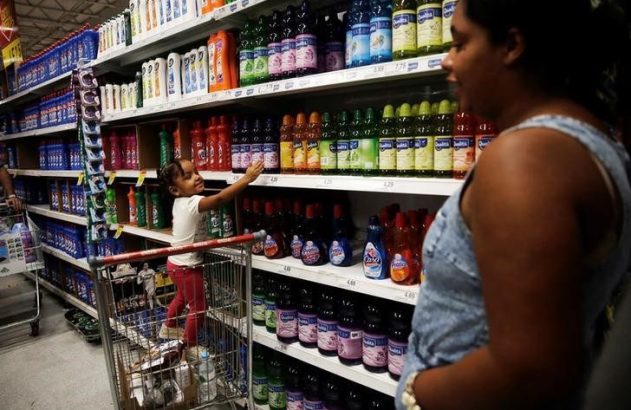 Mãe e filha em supermercado. Criança em carrinho alcança produto de limpeza na prateleira.