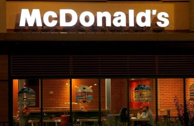 Fachada de McDonald's à noite. Letreiro iluminado, clientes comendo e decorações de hambúrguer nas janelas.