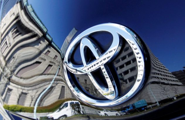 Emblema Toyota cromado em capô escuro, com reflexos distorcidos de prédios e céu.