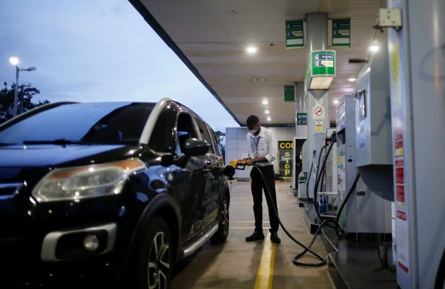 Frentista de máscara abastece carro preto em posto de gasolina ao anoitecer.