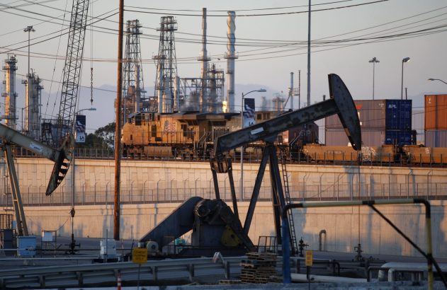 Cavaletes de petróleo e trem de carga com contêineres em frente a uma refinaria industrial ao entardecer.