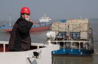 Homem de capacete vermelho e máscara com bandeira da China em um barco, com navios de carga no rio.