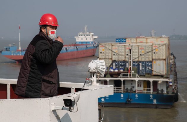 Homem de capacete vermelho e máscara com bandeira da China em um barco, com navios de carga no rio.
