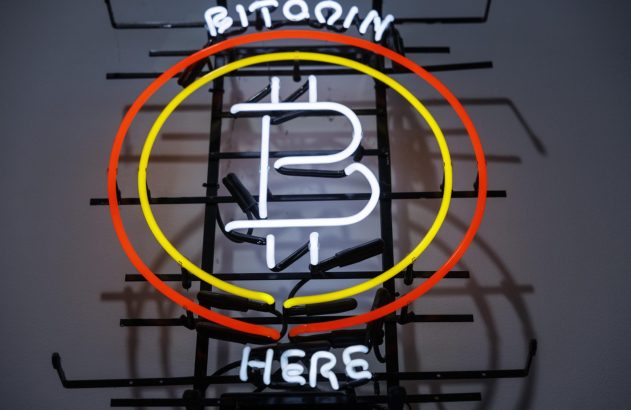 Sinal de neon do Bitcoin: logo 'B' com linhas verticais, cercado por círculos amarelo e laranja. Texto 'BITCOIN HERE' iluminado.