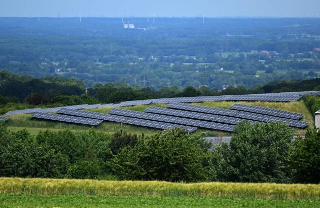 Painéis solares em encosta verde, com plantações, árvores e turbinas eólicas no horizonte nebuloso.