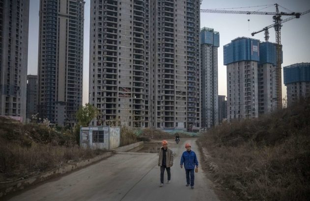 Dois trabalhadores caminham por estrada de terra. Ao fundo, muitos arranha-céus inacabados, alguns com andaimes azuis e guindastes.