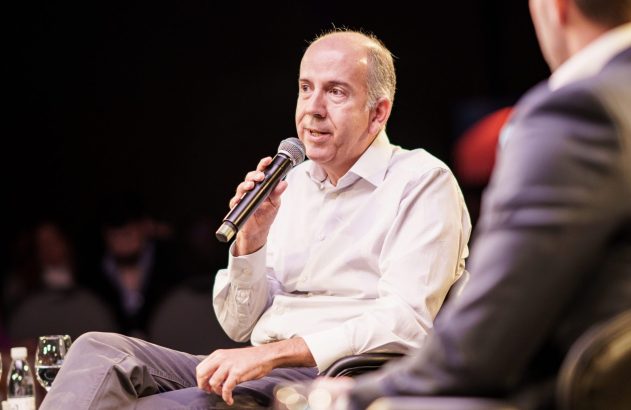 Homem calvo de camisa clara sentado, falando em microfone. As costas de outra pessoa estão visíveis à direita.