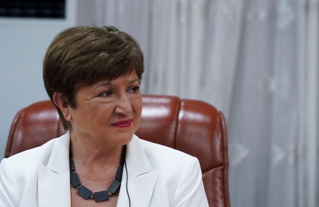 Mulher sentada em blazer branco e colar preto, com cabelo curto, sorrindo suavemente e olhando para a esquerda.