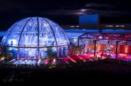 Evento noturno com cúpula transparente azul e lounge externo vermelho, com pessoas e plantas.