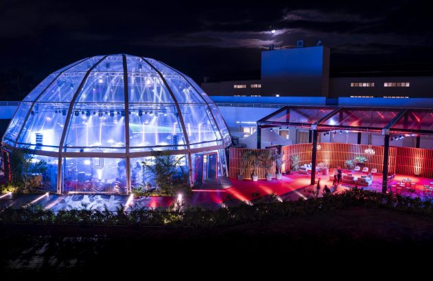 Evento noturno com cúpula transparente azul e lounge externo vermelho, com pessoas e plantas.