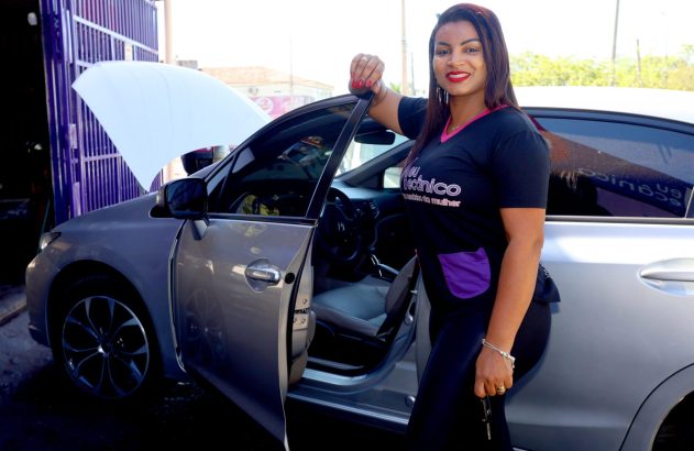 Mulher sorrindo ao lado de carro prateado com capô e porta abertos, segurando chaves.