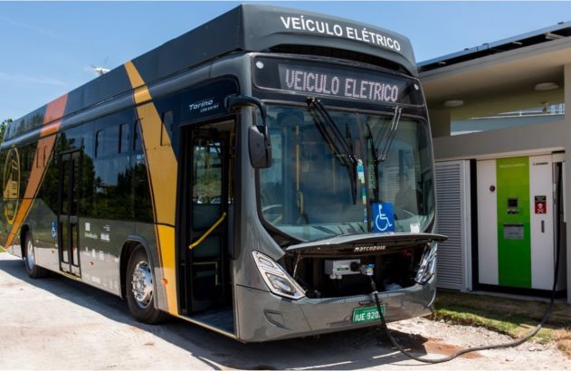 Ônibus elétrico cinza e amarelo sendo carregado em uma estação de recarga rápida.